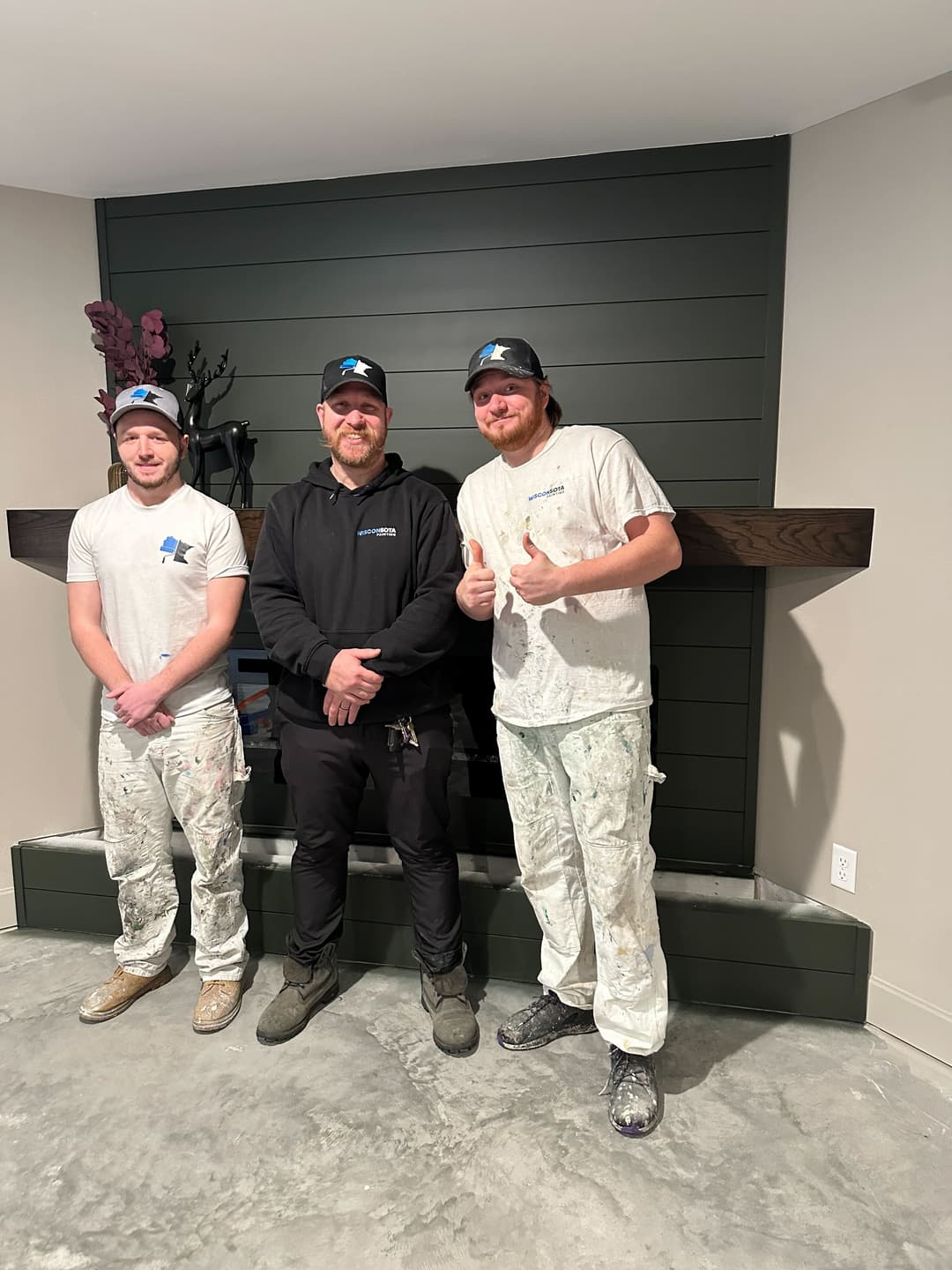 Three painters in work attire stand in front of a green accent wall and modern fireplace.