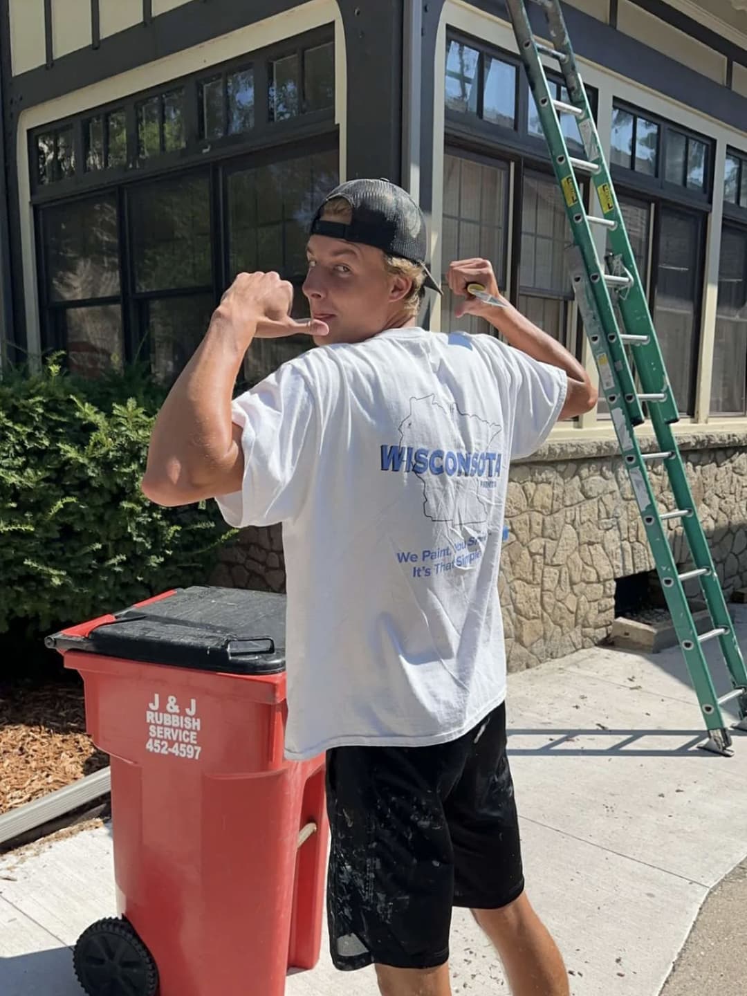 Young man in a Wisconsin shirt flexing muscles by a trash can and ladder on a sunny day.