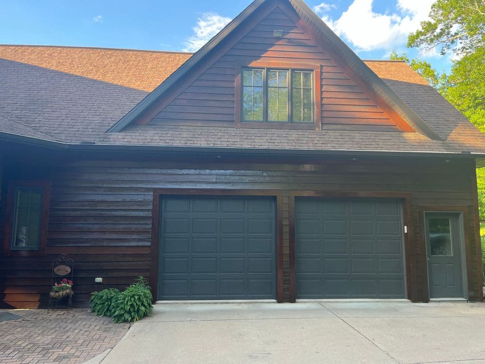 Modern rustic home exterior with dark wood siding and two garage doors. Bright sky above.