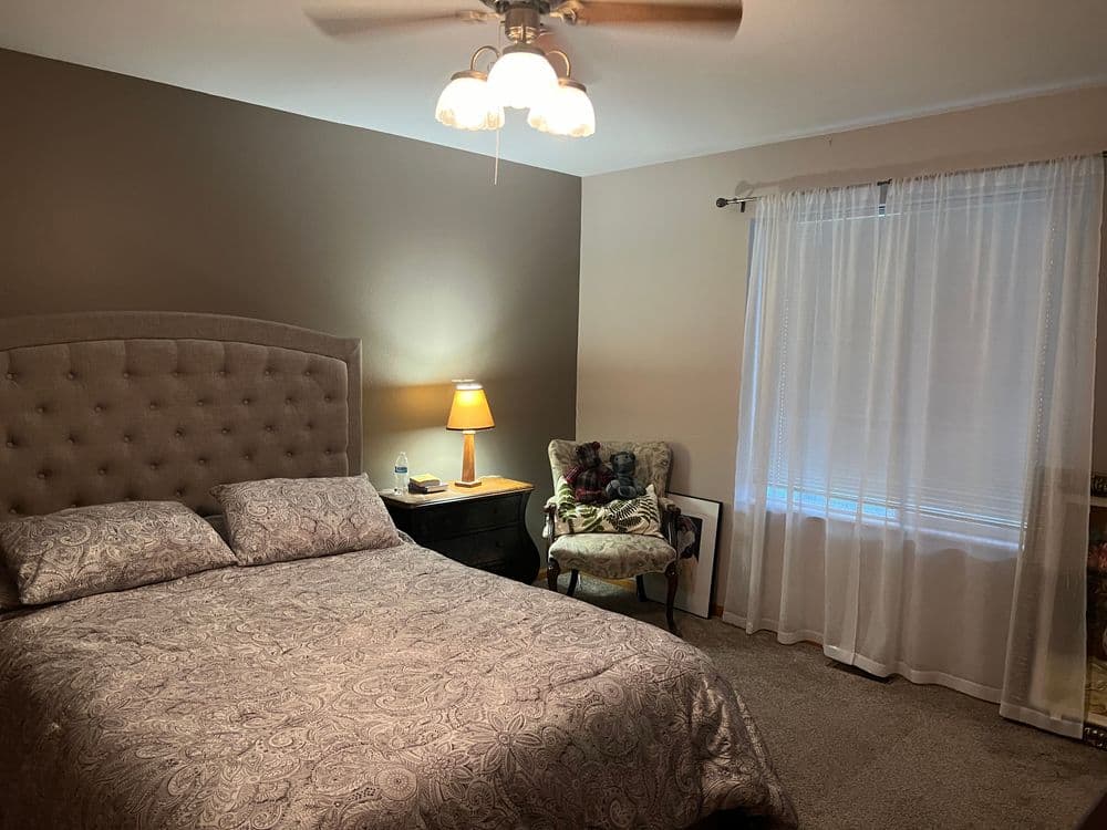 Cozy bedroom with a tufted bed, wooden nightstand, lamp, and sunlight through sheer curtains.