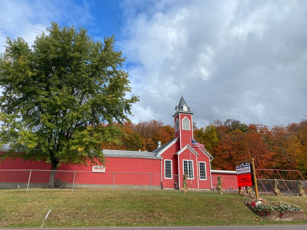 Red historic schoolhouse with a clock tower, surrounded by autumn foliage and a sign.