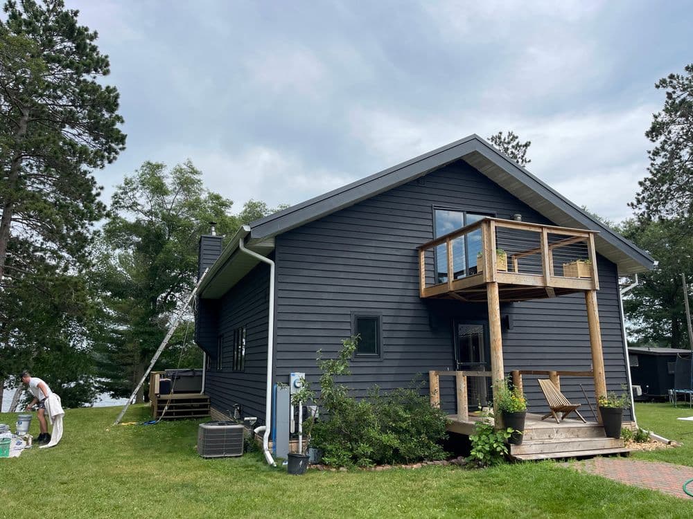 Modern dark gray house with wooden porch, surrounded by trees and lawn by the lake.
