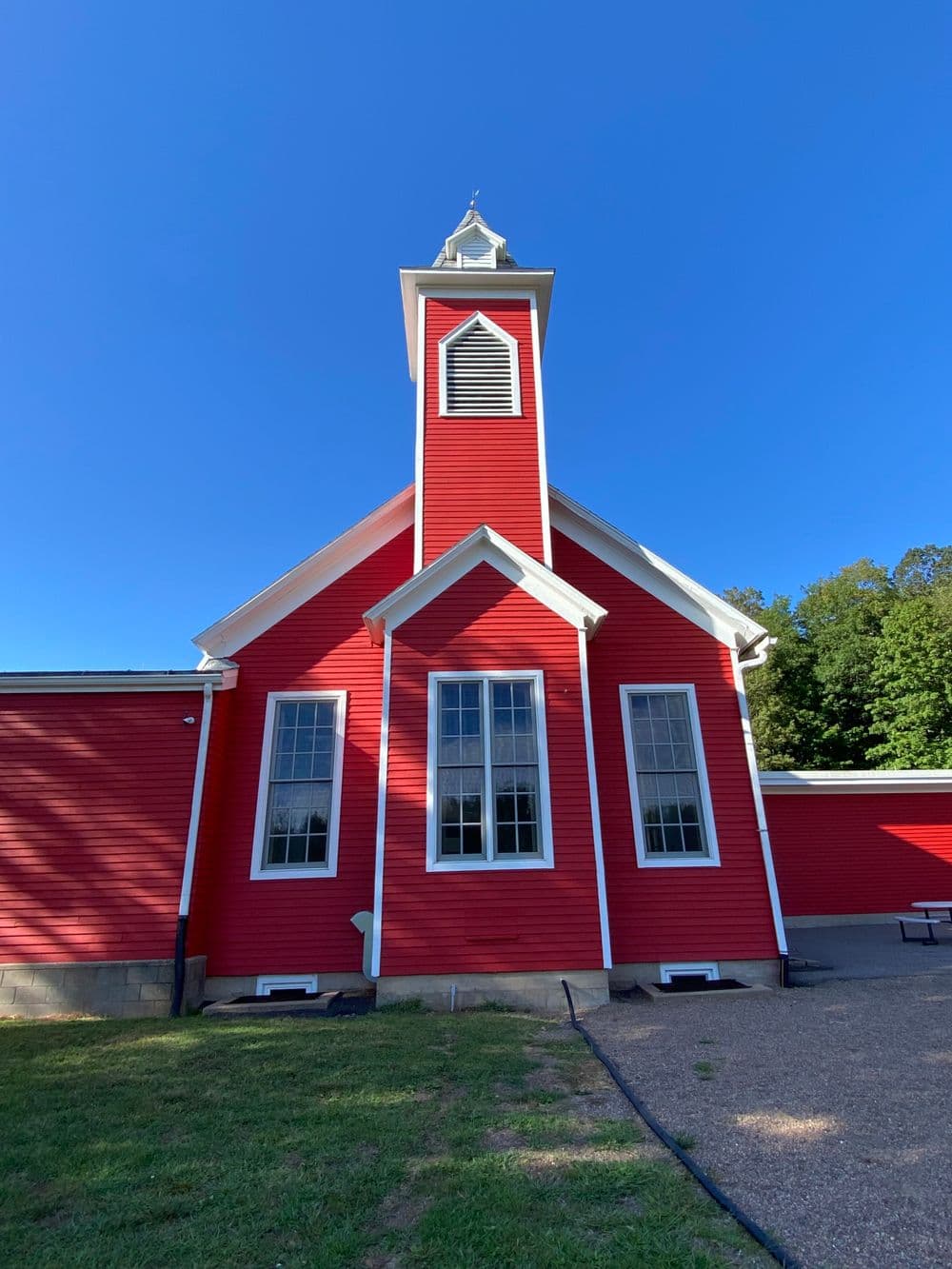 Red church building with a tall steeple against a clear blue sky. Green trees in the background.