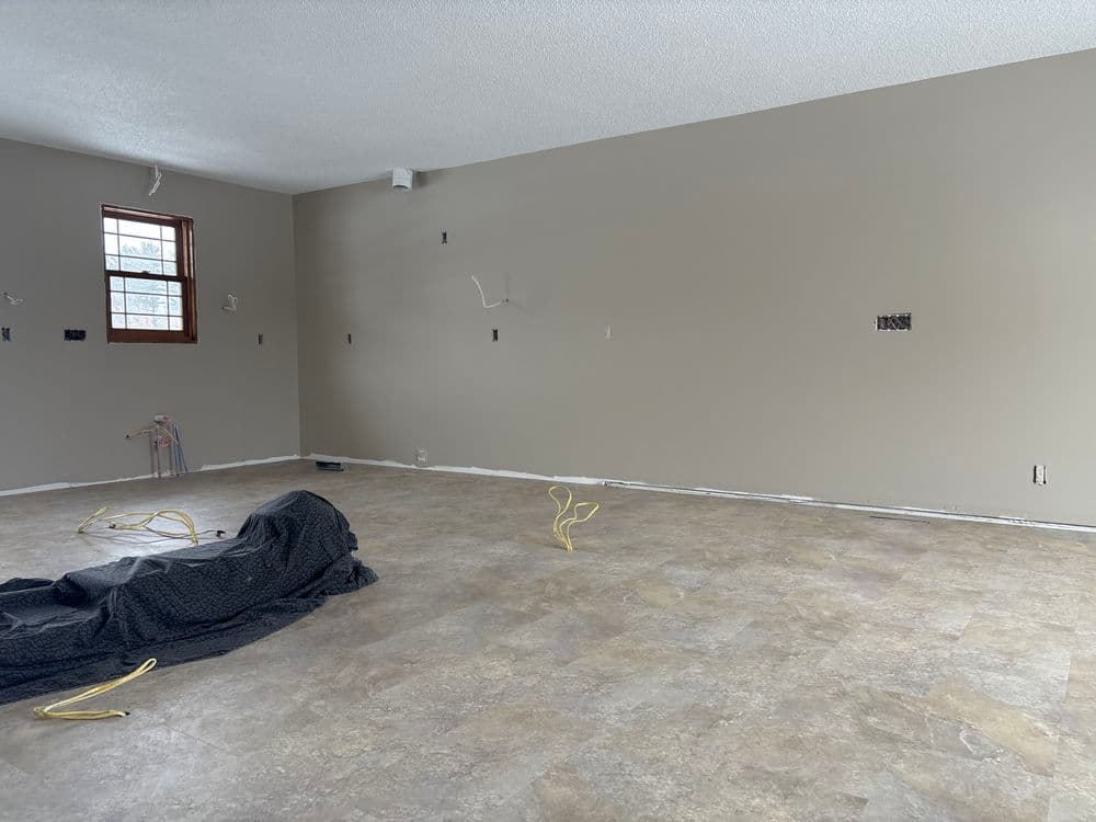 Empty, renovated living room with beige walls and unfinished flooring, featuring cables and a black tarp.