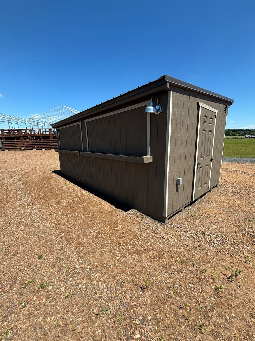 Side view of a brown shed with a door and wall-mounted lamp against a blue sky.