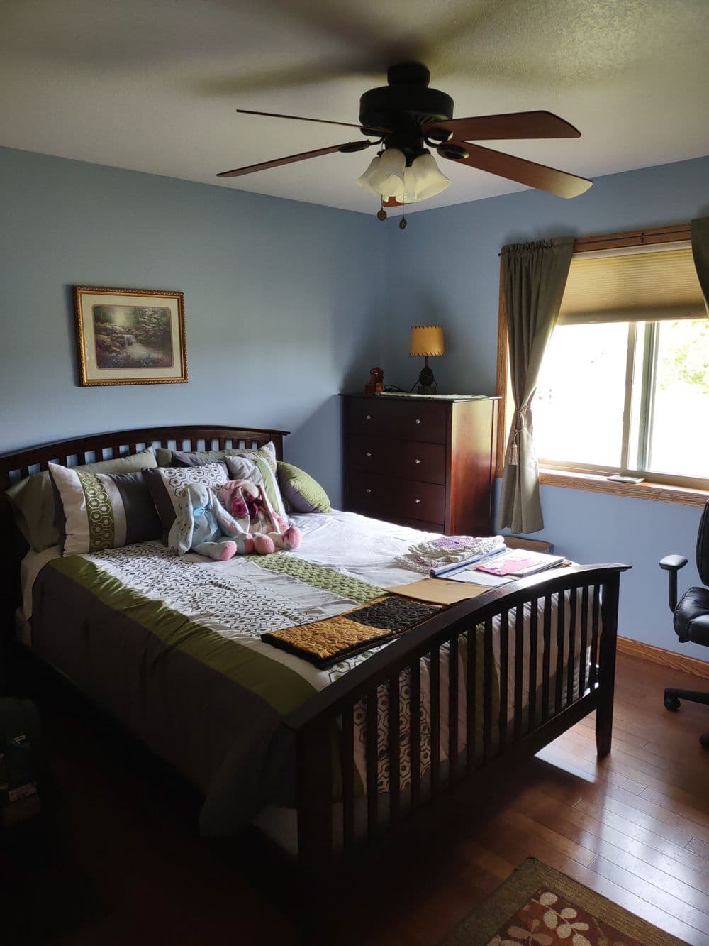 Cozy bedroom with a blue wall, wooden bed, ceiling fan, and a window with natural light.