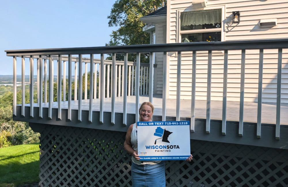 Woman holding a Wisconsin Painting sign in front of a newly painted deck with a scenic view.