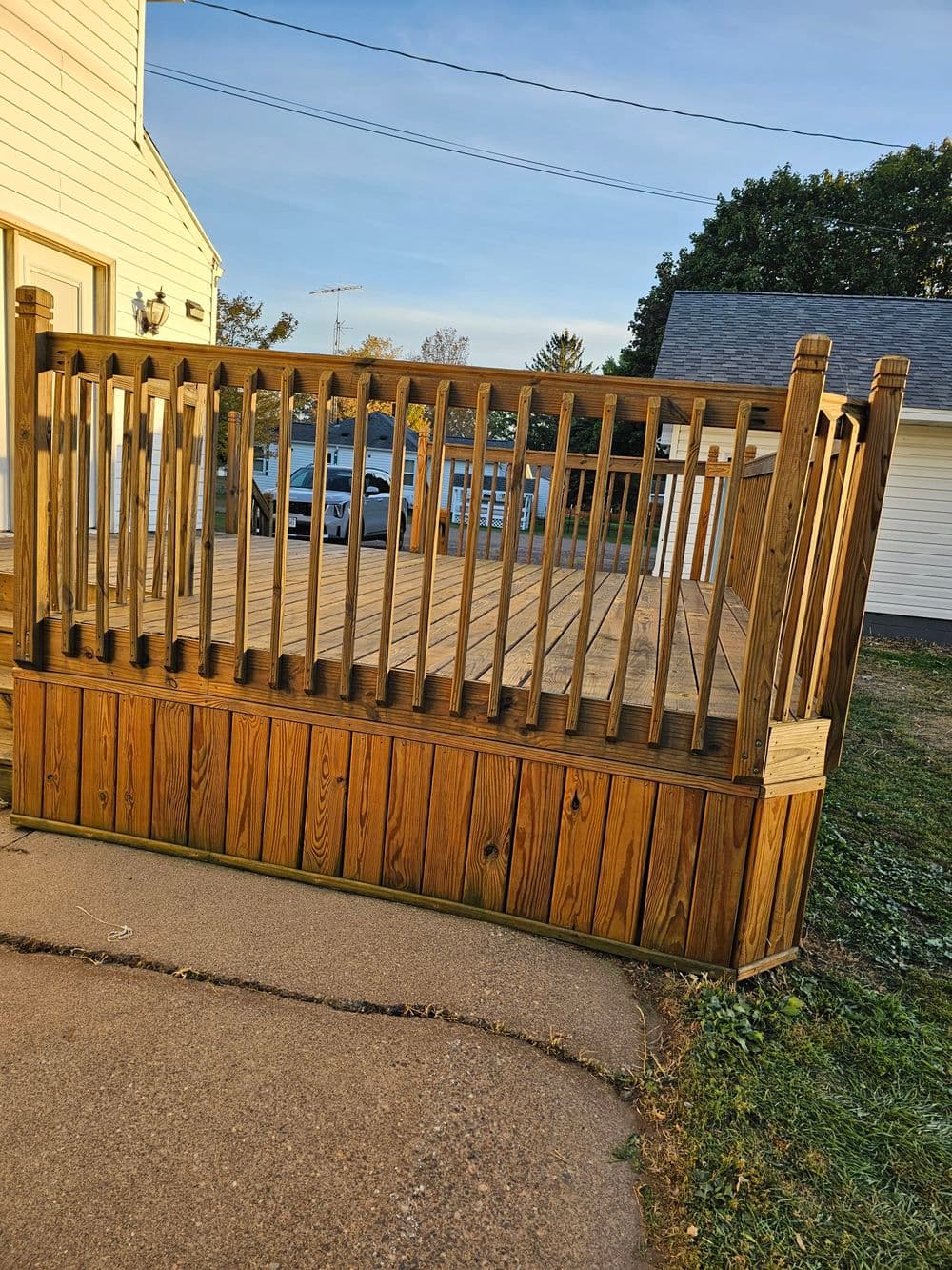 Wooden deck with railing, grass and concrete path, residential setting and clear sky.