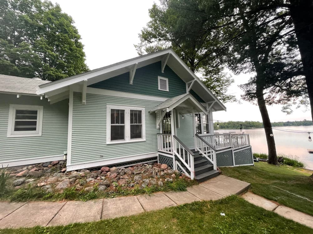 Charming lakeside cottage with green siding, porch, and stone landscaping by the water.