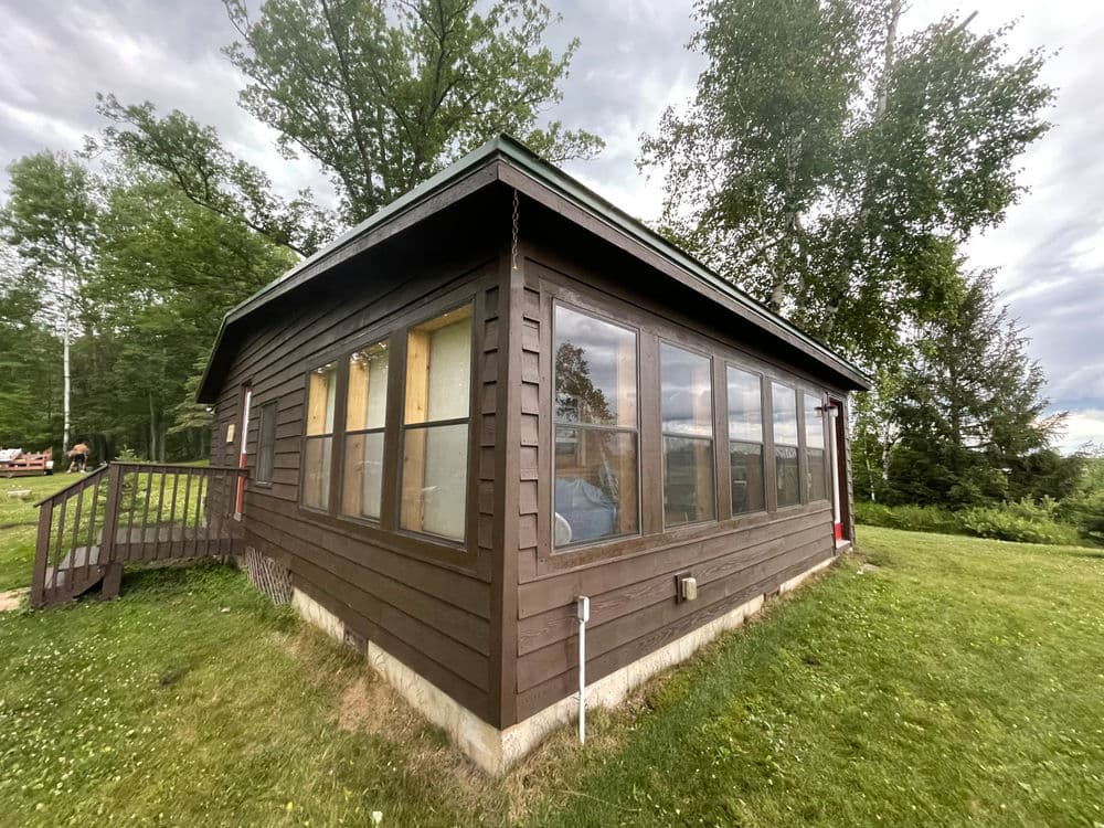 Rustic brown cabin with large windows surrounded by lush greenery and trees.