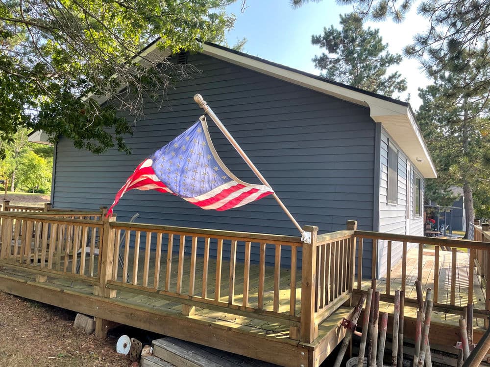 American flag waving on a wooden deck by a blue house surrounded by trees.