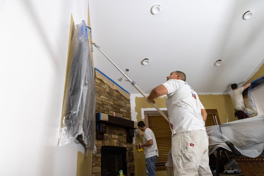 Two professional painters applying fresh paint in a living room with a stone fireplace.