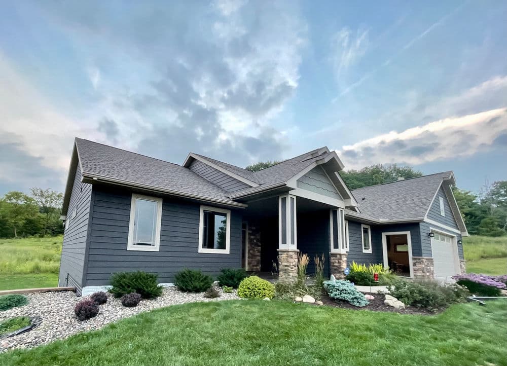 Modern gray house with stone accents, lush landscaping, and a cloudy sky backdrop.