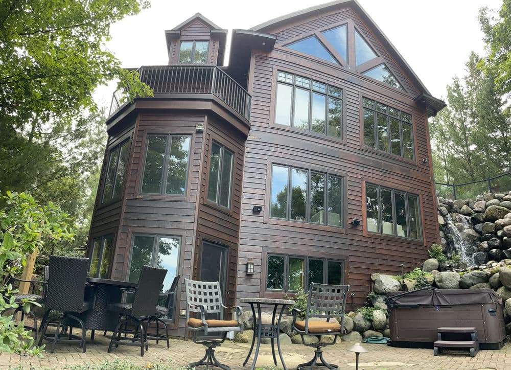 Modern wooden lakeside house with large windows, patio furniture, and stone waterfall feature.