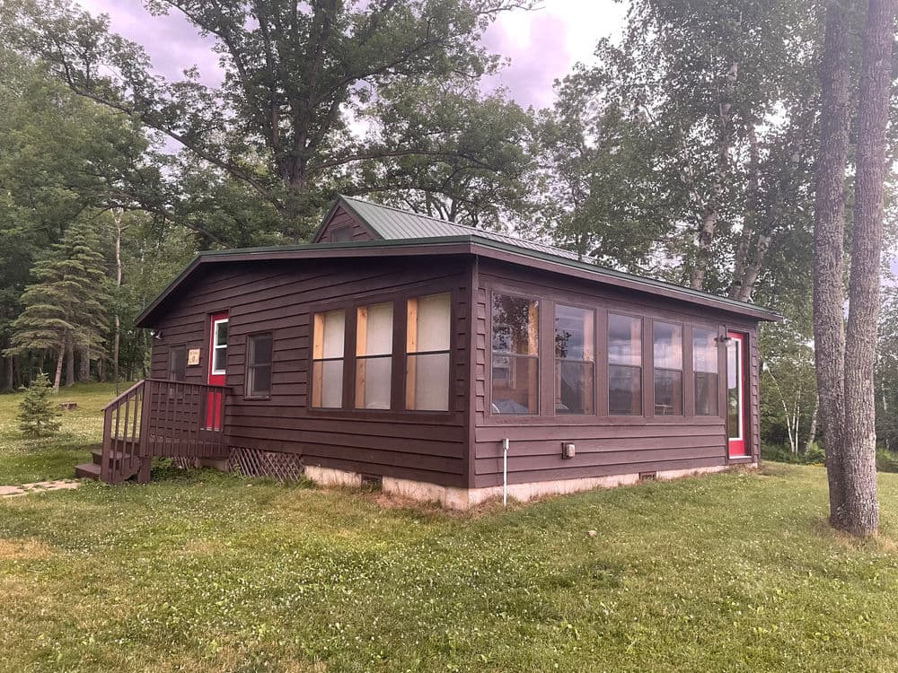 Cozy cabin with screened porch surrounded by lush greenery and tall trees.